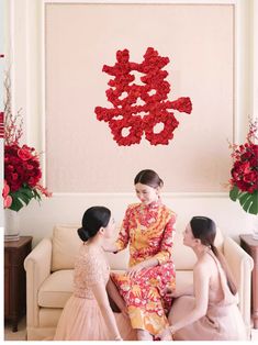 three women sitting on a couch in front of a wall with red flowers hanging from it