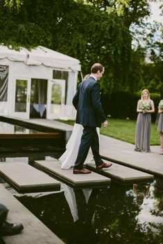 a man in a suit and tie walking across a wooden platform over water with other people