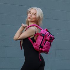 a woman with blonde hair wearing a pink backpack and black leggings stands in front of a gray brick wall