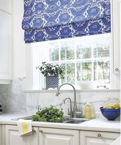 a kitchen with white cabinets and blue patterned roman shades on the window over the sink