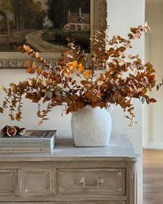 a white vase filled with lots of leaves on top of a wooden dresser next to a painting