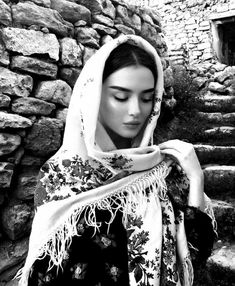 a woman wearing a shawl standing in front of a stone wall with her eyes closed