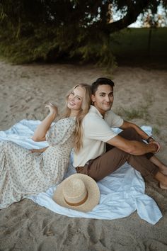 a man and woman sitting on the sand with a hat in their hands, smiling at the camera