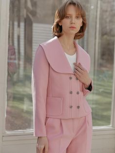 a woman standing in front of a window wearing a pink suit and white shirt with buttons