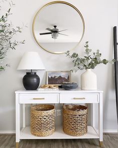 a white table with two baskets under a round mirror on the wall next to a lamp