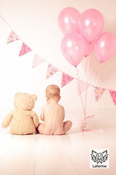 a baby sitting next to a teddy bear with pink and white balloons in the air