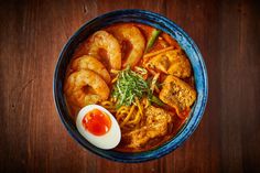 a blue bowl filled with soup and shrimp next to an egg on top of a wooden table