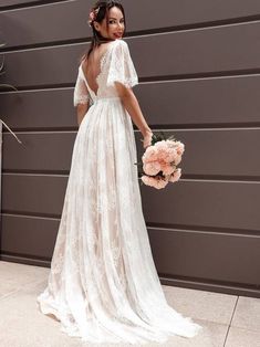 a woman standing in front of a wall wearing a white dress and holding a bouquet