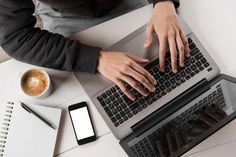 a person typing on a laptop computer next to a cup of coffee and cell phone