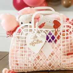 a teddy bear sitting in a white basket filled with pink and white stuff animals next to balloons