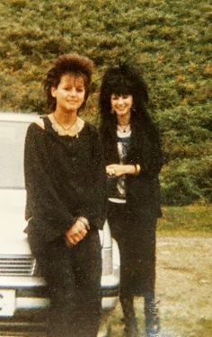 two women standing next to each other in front of a white car with bushes behind them