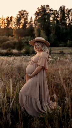 a pregnant woman in a dress and hat standing in tall grass