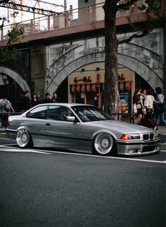 a silver car is parked on the side of the road in front of people and trees