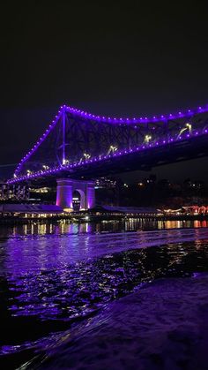 a bridge lit up at night with purple lights on it's sides and water below