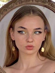 a woman with blue eyes and gold make - up looks into the camera in front of a mirror