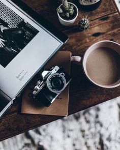 an open book, camera and cup of coffee sit on a table next to cacti