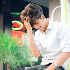 a young man sitting on top of a wooden bench next to a green plant in front of a building