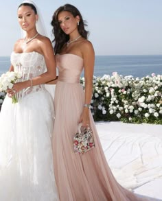 two beautiful women standing next to each other in front of flowers and bushes with the ocean in the background