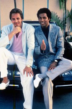 two men sitting on the back of a car posing for a photo in front of a building