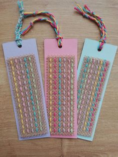 three bookmarks with colorful thread on them sitting on a wooden table next to each other