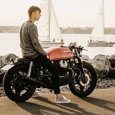 a man sitting on a red motorcycle next to the ocean and sailboats in the background