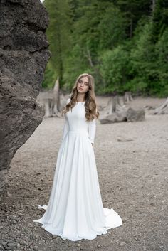 a woman in a white wedding dress standing next to a large rock and looking at the camera