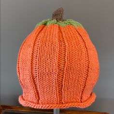 an orange knitted pumpkin hat sitting on top of a wooden stand next to a gray wall