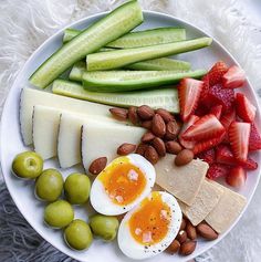 a white plate topped with cheese, fruit and crackers next to cucumbers