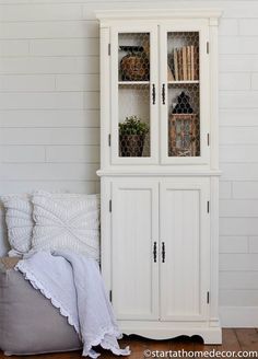 a white china cabinet with glass doors on the top and bottom, next to a bed