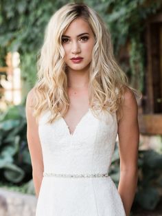 a woman with blonde hair wearing a white dress and looking at the camera while standing in front of greenery