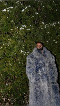 a man in a fur coat standing next to a bush with snow on the ground