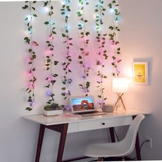 a white desk topped with a laptop computer next to a wall covered in vines and lights