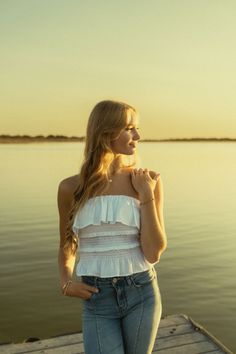 a beautiful young woman standing on top of a wooden pier next to the ocean at sunset