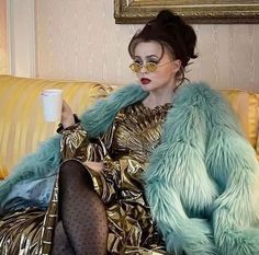 a woman sitting on a couch with a cup in her hand and wearing a fur coat