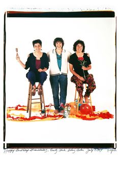 three women sitting on stools in front of a white background with orange and red paint