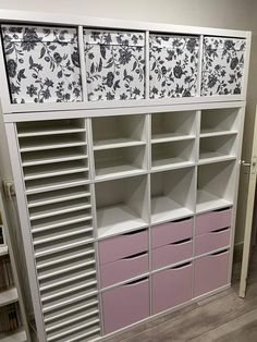 a white bookcase with pink drawers and floral wallpaper on the top, in front of a bookshelf