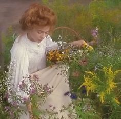 a woman sitting in the grass surrounded by wildflowers and other plants, holding a basket with flowers