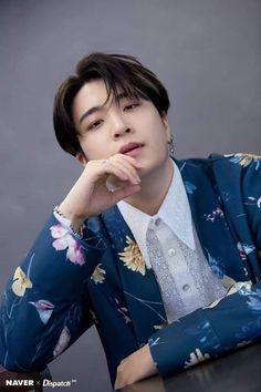 a young man in a blue floral suit and white shirt sitting at a table with his hand on his chin