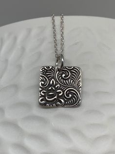 This minimalist tiny rose necklace has been entirely handmade using .999 fine silver. It has been cut out from fine silver precious metal clay, textured, fired and oxidized. The pendant hangs from a sterling silver diamond cut cable chain. This fine silver pendant features an embossed floral rose textured pattern on one side of the pendant and is reversible to a completely different embossed rose pattern on the reverse side. Pendant size: approximately 1/2 x 1/2 inches Chain length: adjustable a Unique Etched Sterling Silver Necklace, Unique Hand Stamped Sterling Silver Jewelry, Etched Sterling Silver Necklaces, Silver Etched Sterling Silver Necklace, Artisan Silver Etched Necklace, Antique Silver Etched Sterling Silver Necklace, Silver Etched Square Pendant Necklace, Sterling Silver Etched Necklace For Gift, Etched Sterling Silver Necklace For Gift