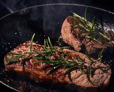 two steaks cooking on a grill with rosemary sprigs in the skillet