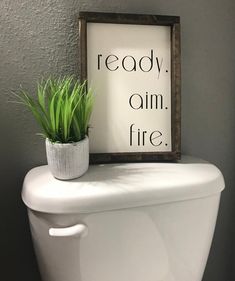 a toilet with a sign that says ready, aim, fire next to it is a potted plant