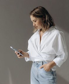 a woman in white shirt and jeans holding a cell phone with her right hand on her hip