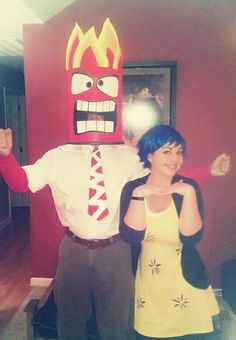 a man and woman in costume standing next to each other near a red wall with a large mask on it's head