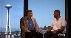 two men sitting next to each other in front of a window with the space needle in the background