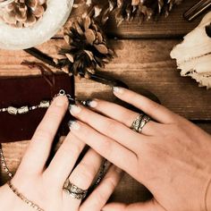 two hands holding each other on a table with jewelry and pine cones in the background