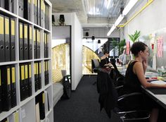 two people sitting at a desk in an office with yellow and black binders on the wall