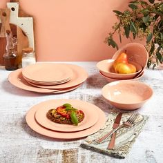 a table topped with pink plates and bowls filled with food next to a vase full of fruit