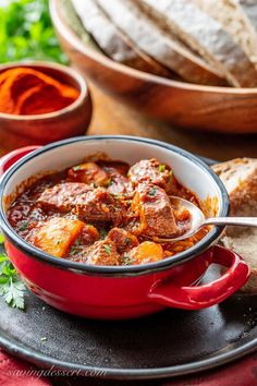 a red bowl filled with meat and vegetables next to bread on a black platter