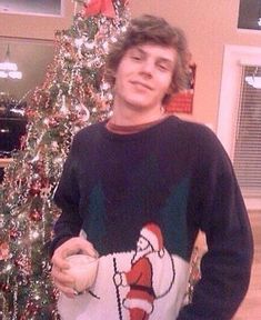 a man standing in front of a christmas tree wearing a sweater with santa clause on it