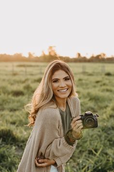 a woman standing in a field holding a camera and smiling at the camera with her hands on her hips